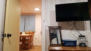 A room with a wooden ceiling, a television mounted on the wall, a table with chairs, a mirror, and a box labeled "Less House more home". - Cozy Cottage, Dog Friendly, Close to beach, Natural Wood, Waterfall on site (Barton)