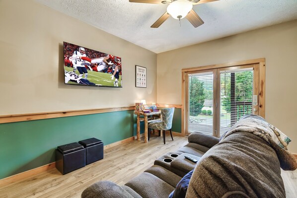 Une pièce avec un ventilateur de plafond, un téléviseur diffusant un match de football, un trophée encadré, une petite table avec une lampe et une fenêtre avec des stores.
