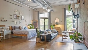 A spacious room with a bed, a sofa, and a small table. There are plants and a window providing natural light.