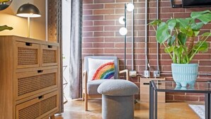 A cozy room with a brick wall, a wooden cabinet, a chair with a rainbow pillow, a round ottoman, and a potted plant.