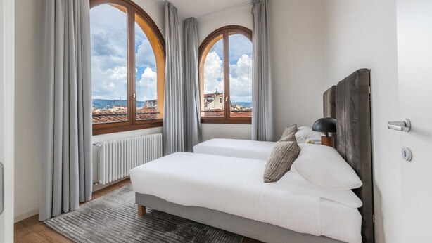 Une chambre à coucher avec un lit, deux fenêtres, un radiateur et une vue sur des immeubles.
