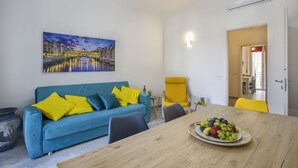 Ein Wohnzimmer mit einem blauen Sofa, gelben Kissen, einem Esstisch mit Obst, einem Stadtbild als Gemälde und einem leuchtend gelben Stuhl.