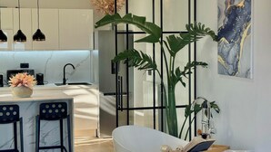 A modern kitchen with white cabinets, a marble countertop, and pendant lighting. There is a large plant and a bathtub with a wooden tray and books. - Casa Benita (Hvar)