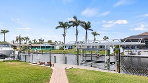 A waterfront walkway with a boat dock, palm trees, and a pedestrian bridge. - Waterfront Oasis, Pool, Dock, Location! (Cape Coral)