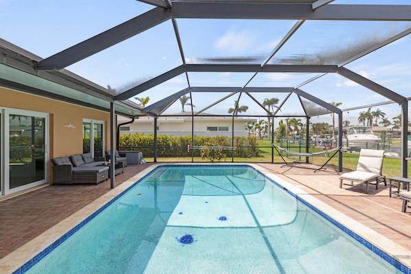 A covered swimming pool with a glass enclosure, outdoor seating, and a view of palm trees and a boat in the background.