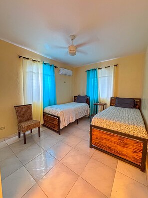 A room with two single beds, a chair, a small table, and an air conditioning unit.