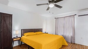 Un dormitorio con cama, ventilador de techo, mesita de noche con lámpara, armario y ventana con cortinas.