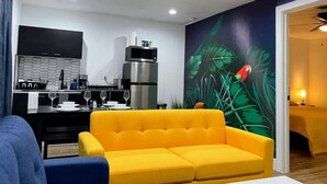 Una sala de estar moderna con un sofá amarillo brillante, una zona de cocina con salpicadero blanco y negro, y un mural de un loro en la pared.