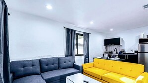 Una sala moderna con un sofá gris y un sofá amarillo, una mesa de centro blanca y una zona de cocina con un armario negro y un refrigerador de acero inoxidable.