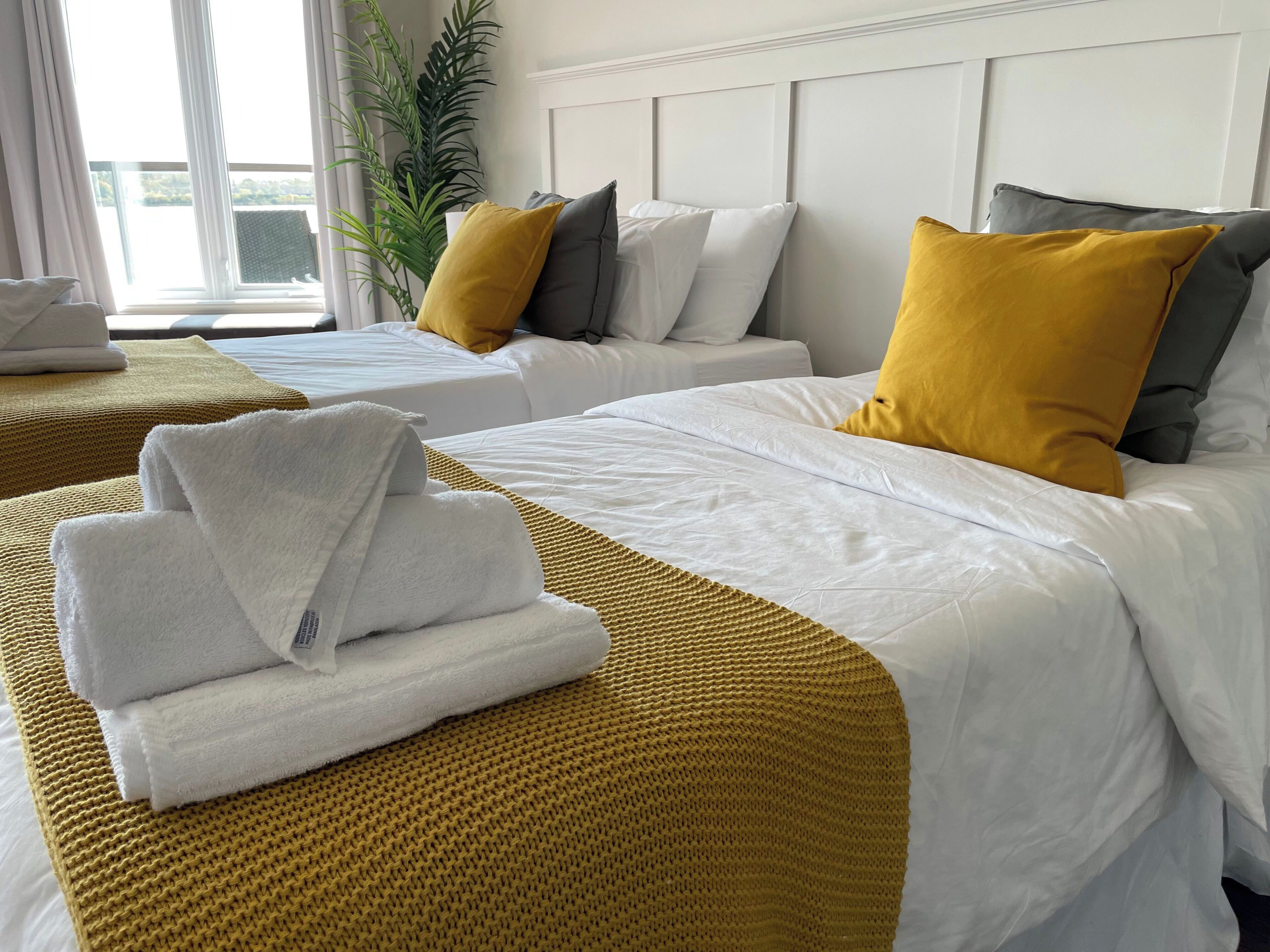 Habitación de hotel con dos camas, ropa de cama blanca, almohadones color mostaza y un ventanal con vistas.