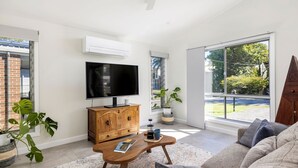 Un salon moderne doté d’un téléviseur à écran plat, d’un meuble en bois, d’une table basse et de grandes fenêtres offrant une vue sur la verdure et un bâtiment en briques.