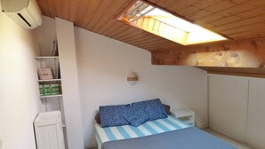 Une petite chambre avec un lit simple, un plafond en pente et un puits de lumière. On y trouve un radiateur blanc, une petite table de chevet blanche et une étagère avec des livres et des boîtes.