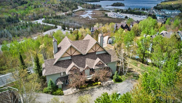 A large house with a steeply pitched roof, surrounded by trees and a lake in the background. - Chalumeau - RVMT (Mont-Tremblant)