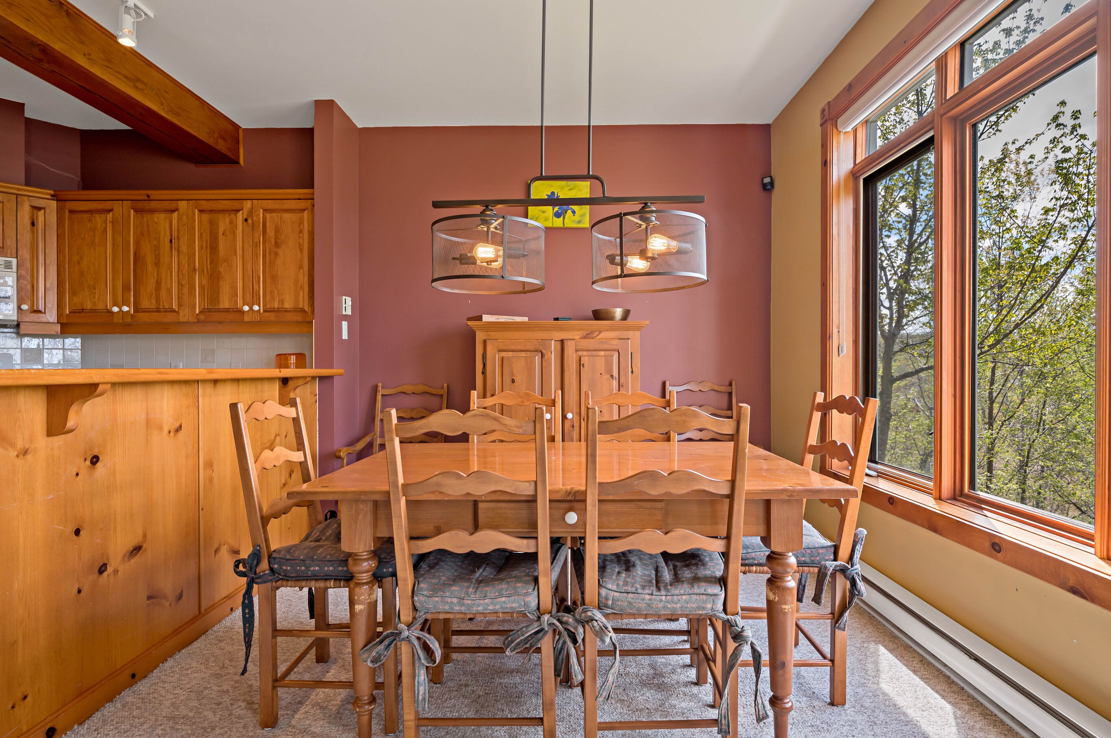 A dining area with a wooden table and chairs, a wooden cabinet, and a window with a view of trees.