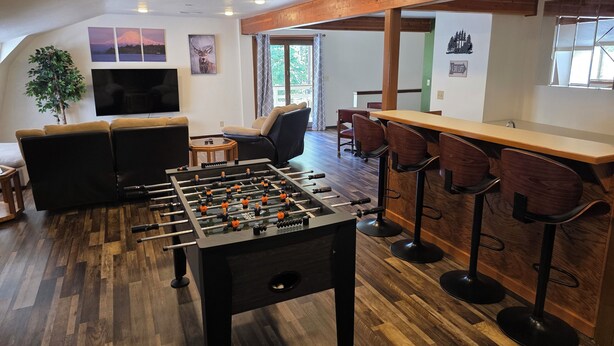 A cozy living room with a foosball table, a sofa, and a TV. There are wooden floors and a bar area with stools.