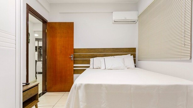 Une chambre à coucher avec un lit, une tête de lit en bois, un mur blanc, une fenêtre avec des stores et un appareil de climatisation.