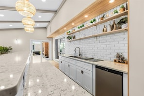 A modern kitchen with white brick backsplash, wooden shelves with plants, and a built-in dishwasher.