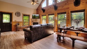 A spacious living room with a fireplace, wooden floors, a leather sofa, and large windows offering a view of the outdoors.