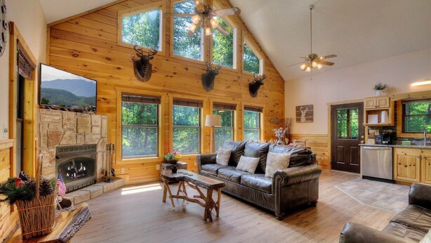 A cozy cabin interior with a stone fireplace, leather sofa, wooden dining table, and a kitchen area.