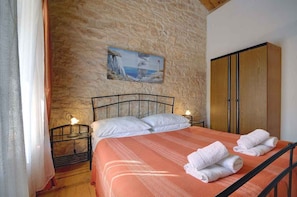 A bedroom with a stone wall, a bed with white bedding, a painting, and a wooden wardrobe.
