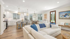 Een moderne keuken met witte kasten, een eethoek met rieten stoelen, en een woonkamer met een beige bank en decoratieve kussens.