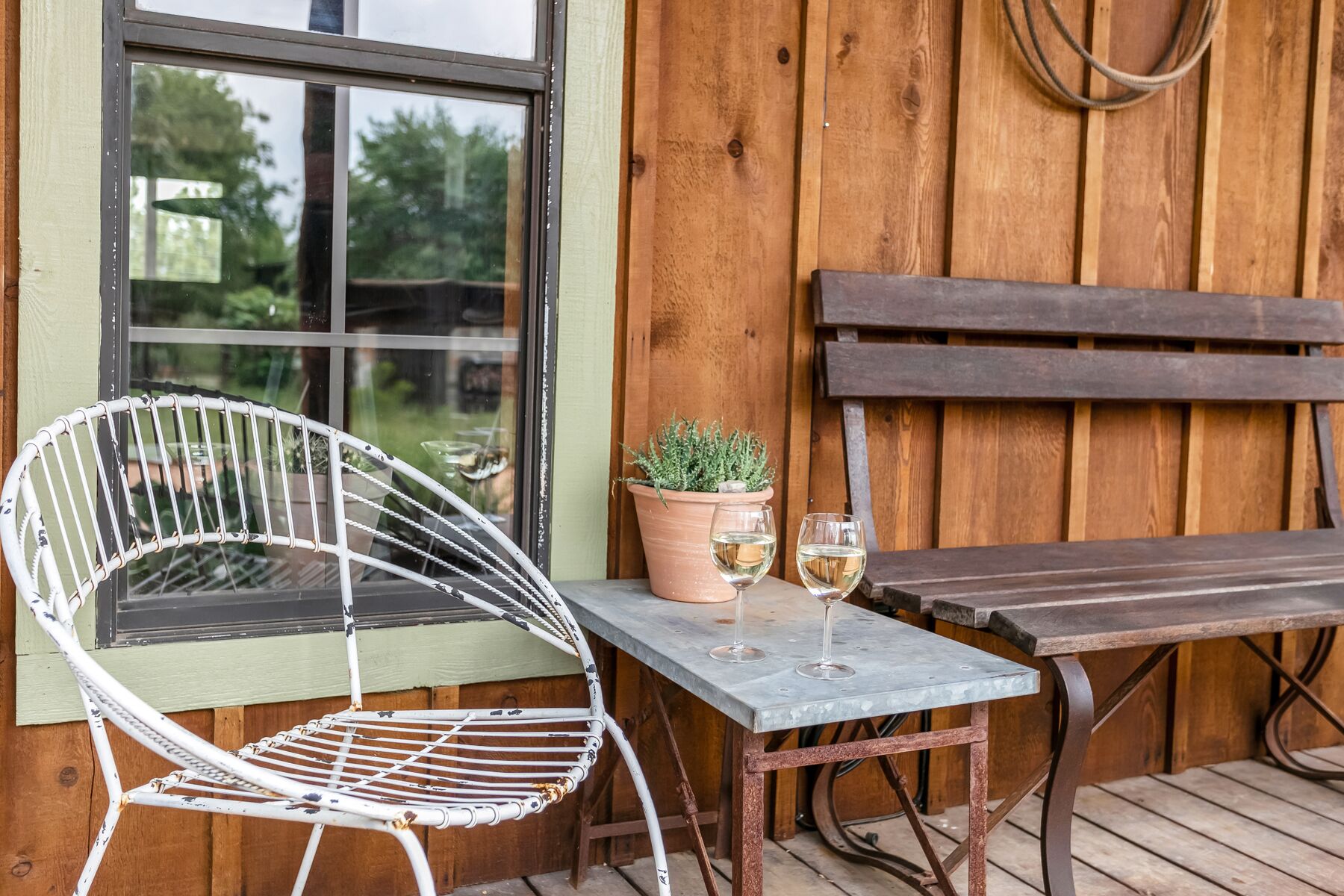 A rustic outdoor seating area with a metal chair, a wooden bench, and a table. There is a potted plant on the table and two glasses of wine.