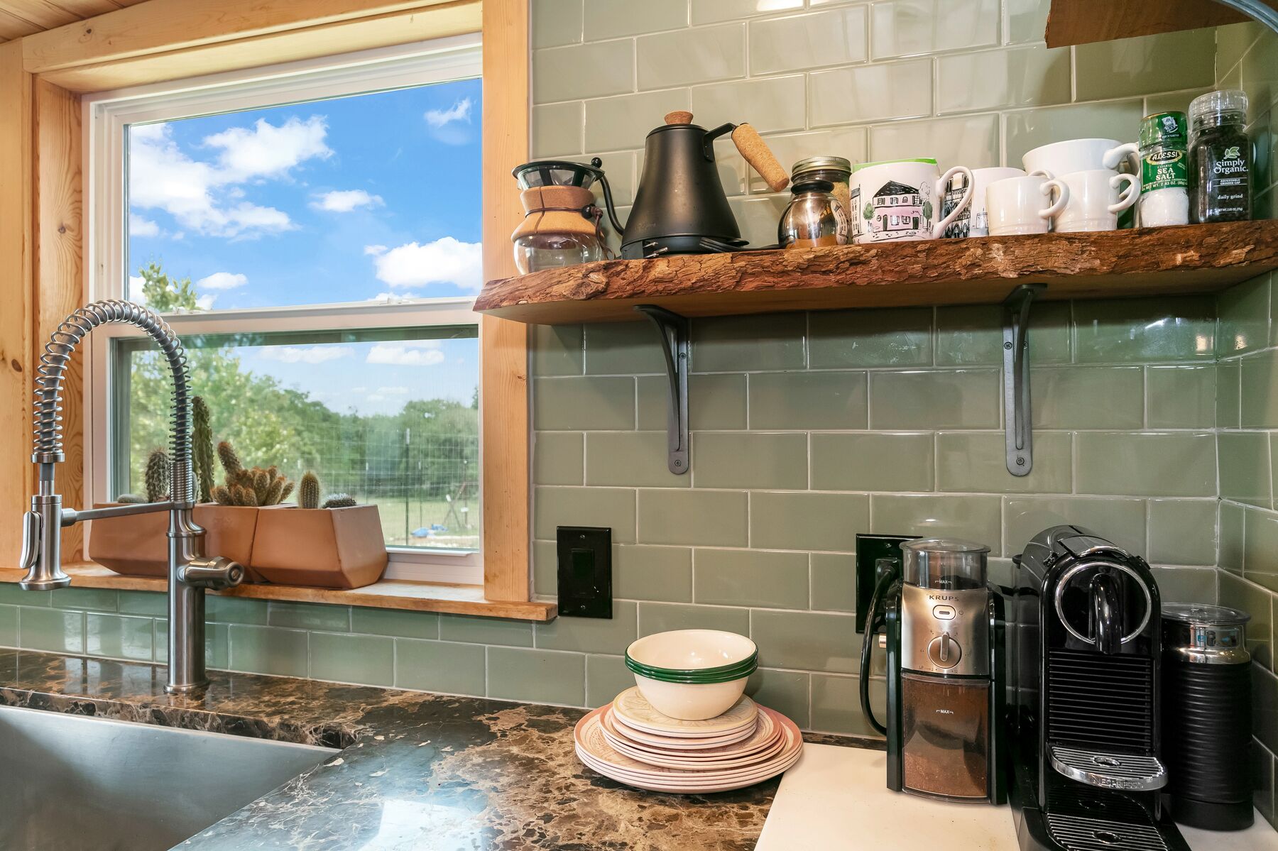Una cocina con ventana, un lavamanos, una cafetera y una estantería con tazas y frascos.