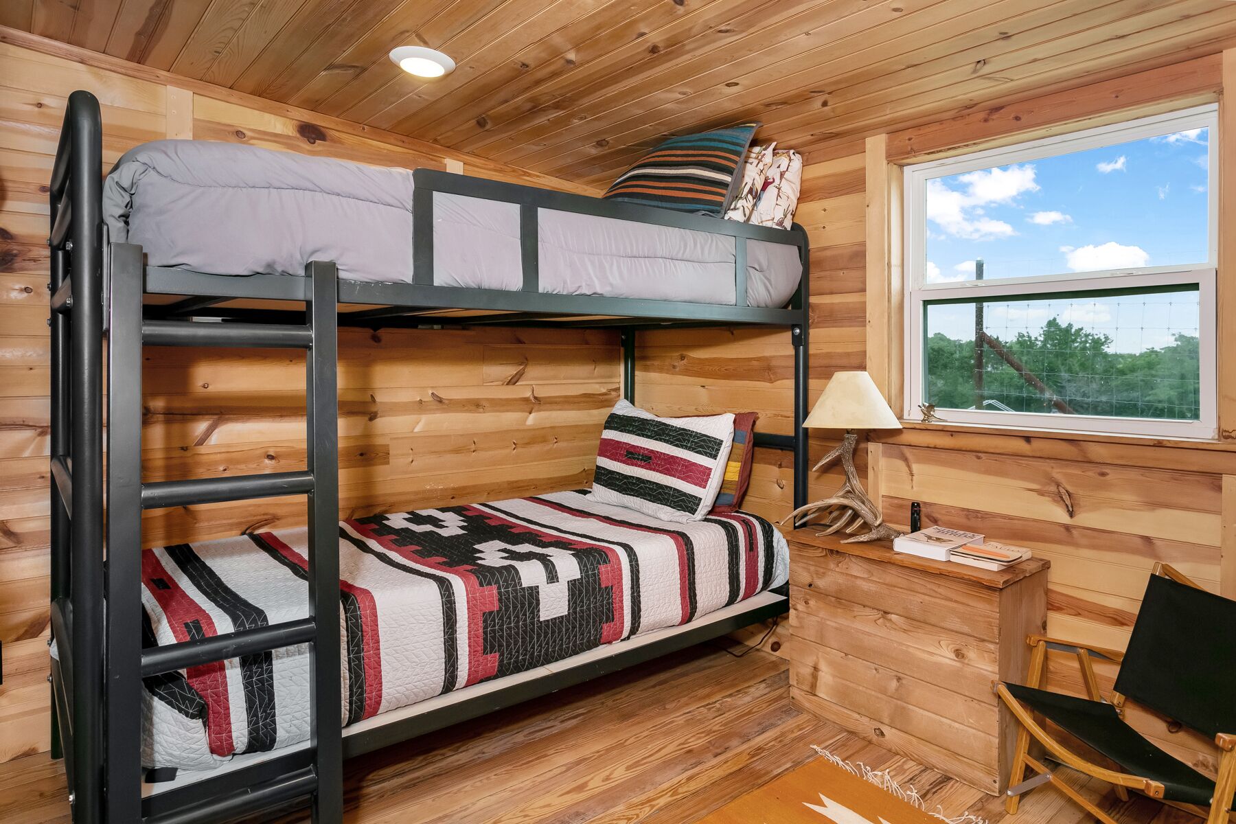 Un lit superposé avec une couverture et des oreillers à motifs, une petite table de chevet en bois avec une lampe, et une fenêtre donnant sur les arbres.