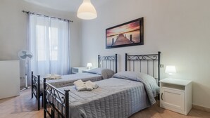 A bedroom with two beds, a bedside table with a lamp, a standing fan, and a painting on the wall.