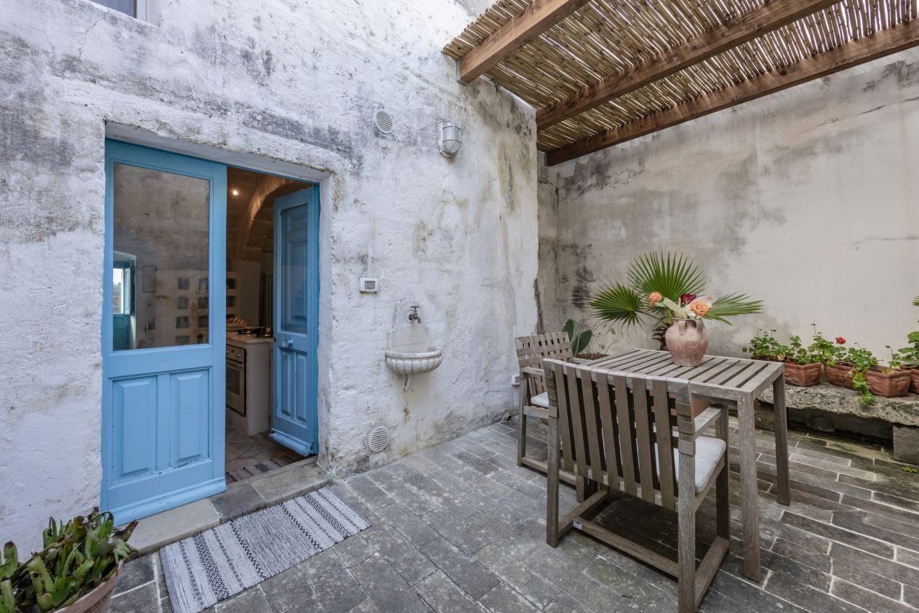 Un espace extérieur rustique avec une table et des chaises en bois, une porte bleue donnant sur une cuisine et des plantes en pot.