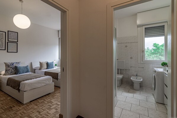 A bedroom with two beds, a large window, and a bathroom with a toilet, bidet, and washing machine.