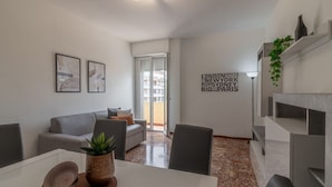 Een moderne woonkamer met een bank, een eettafel met een schaal en plant, een muur met stadsnamen, en een schuifpui.