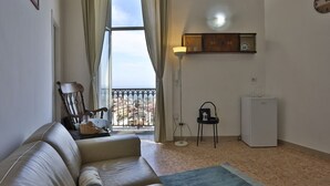 A living room with a sofa, a chair, a small table with a lamp, a refrigerator, and a window with curtains.