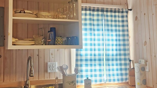 Una cocina con paneles de madera, un estante blanco con cristalería, una cortina de ventana a cuadros y un hervidor blanco sobre la encimera.