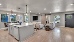 Une cuisine moderne avec un îlot central, un coin repas avec une table et des chaises, un salon avec un canapé et une télévision, et une grande fenêtre donnant sur l’extérieur.