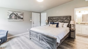 Une chambre à coucher avec un grand lit, une table de chevet avec une lampe, une chaise, un tableau représentant une grange, et une salle de bain avec un miroir et un lavabo.