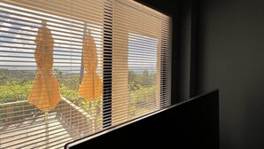 A room with a large window featuring horizontal blinds, a view of greenery and a distant landscape, and a dark wall. - Ocean View Capitol Hill Studio with Sunset Deck (Capitol Hill)