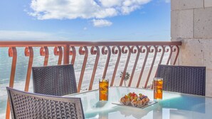 Une table en verre avec deux chaises, une assiette de nourriture et deux boissons sur un balcon donnant sur l’océan.