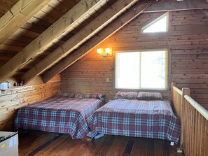 Un dormitorio acogedor en el ático con dos camas, paredes de madera, techo inclinado y una ventana que deja entrar luz natural.