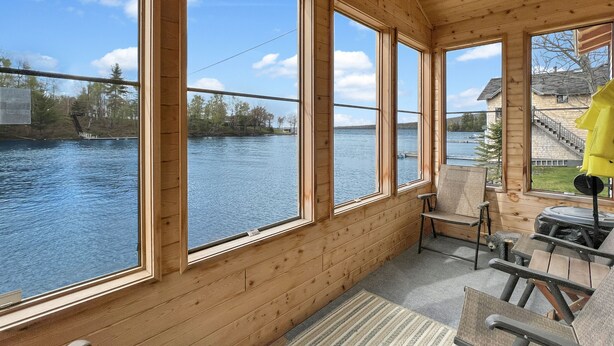 En træhytte med store vinduer ud til en sø, en terrasse med udendørs møbler, og en trappe op til et øvre niveau.