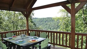 A covered outdoor dining area with a table set for four, surrounded by green chairs, overlooking a forested landscape. - Amazing Views! Hot Tub!! Minutes from the Gorge!!! (Stanton)
