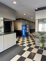 Um saguão com piso quadriculado, uma máquina de refrigerantes Pepsi e uma câmera de segurança no teto.
