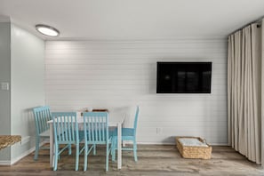 A dining area with a white table and blue chairs, a flat-screen TV mounted on the wall, and a wicker basket on the floor. - Stunning Ocean Oasis: Pools, Views, Great Location (Galveston)