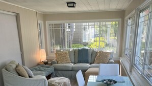 A cozy living room with a sectional sofa, a glass dining table, and a view of the outdoors through large windows. - Cozy Pet Friendly Cottage, Partial Water View, Sleeps 7, Half Block from Beach! (Lexington)