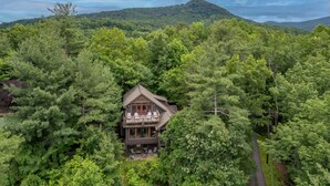 A two-story cabin surrounded by dense forest, with a mountain in the background.