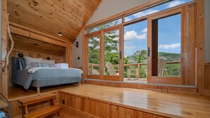 A wooden cabin bedroom with a bed, a bench, and a large window offering a view of the outdoors.