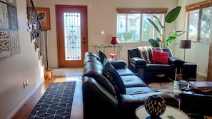 Un salon avec un canapé en cuir noir, une table basse en verre, un vase rouge et une grande fenêtre donnant sur une maison.