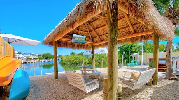 Un área de descanso junto a la playa con techo de paja, mobiliario y vista a la piscina.