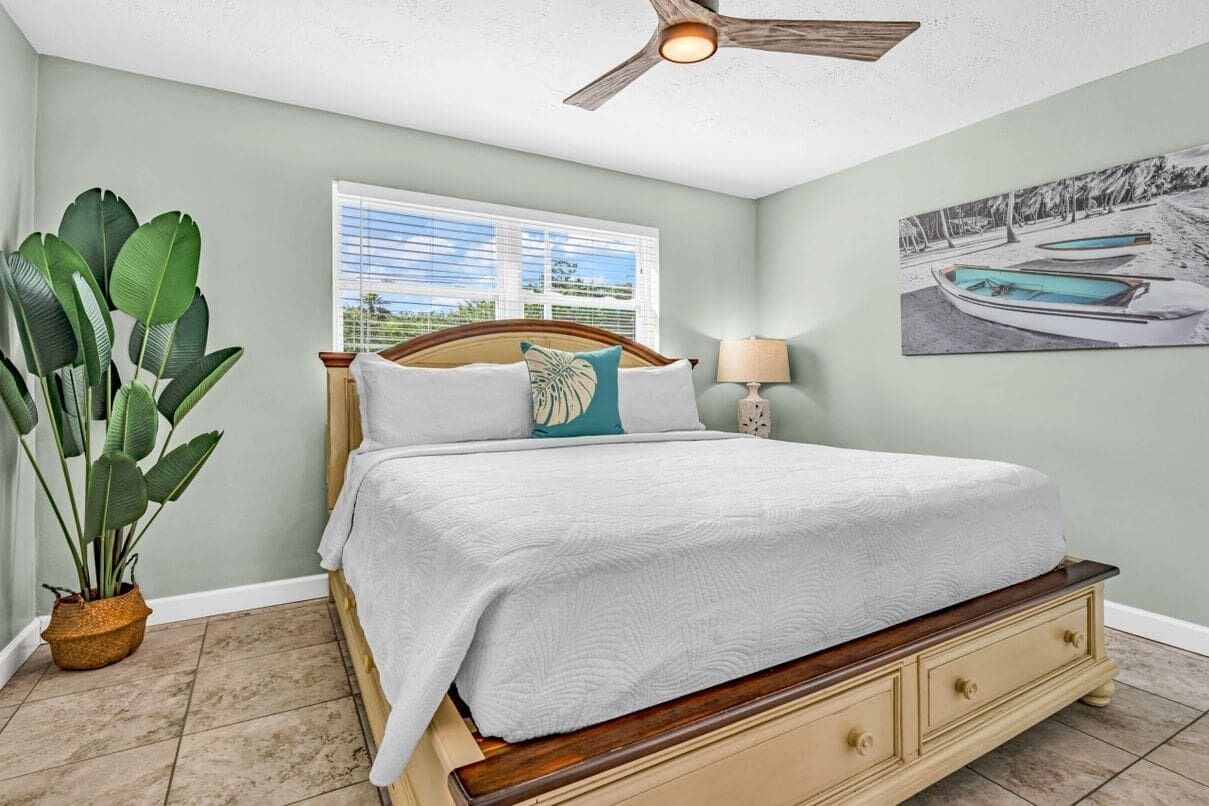 A bedroom with a bed, a ceiling fan, a window with blinds, a potted plant, and a painting on the wall.