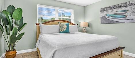 A bedroom with a bed, a ceiling fan, a window with blinds, a potted plant, and a painting on the wall.